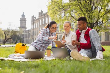 A group of students sit outside their university building on blankets, collaboratively working on a project.