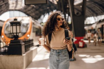 Stylish african woman walks through railway station with smartphone and cup of coffee.