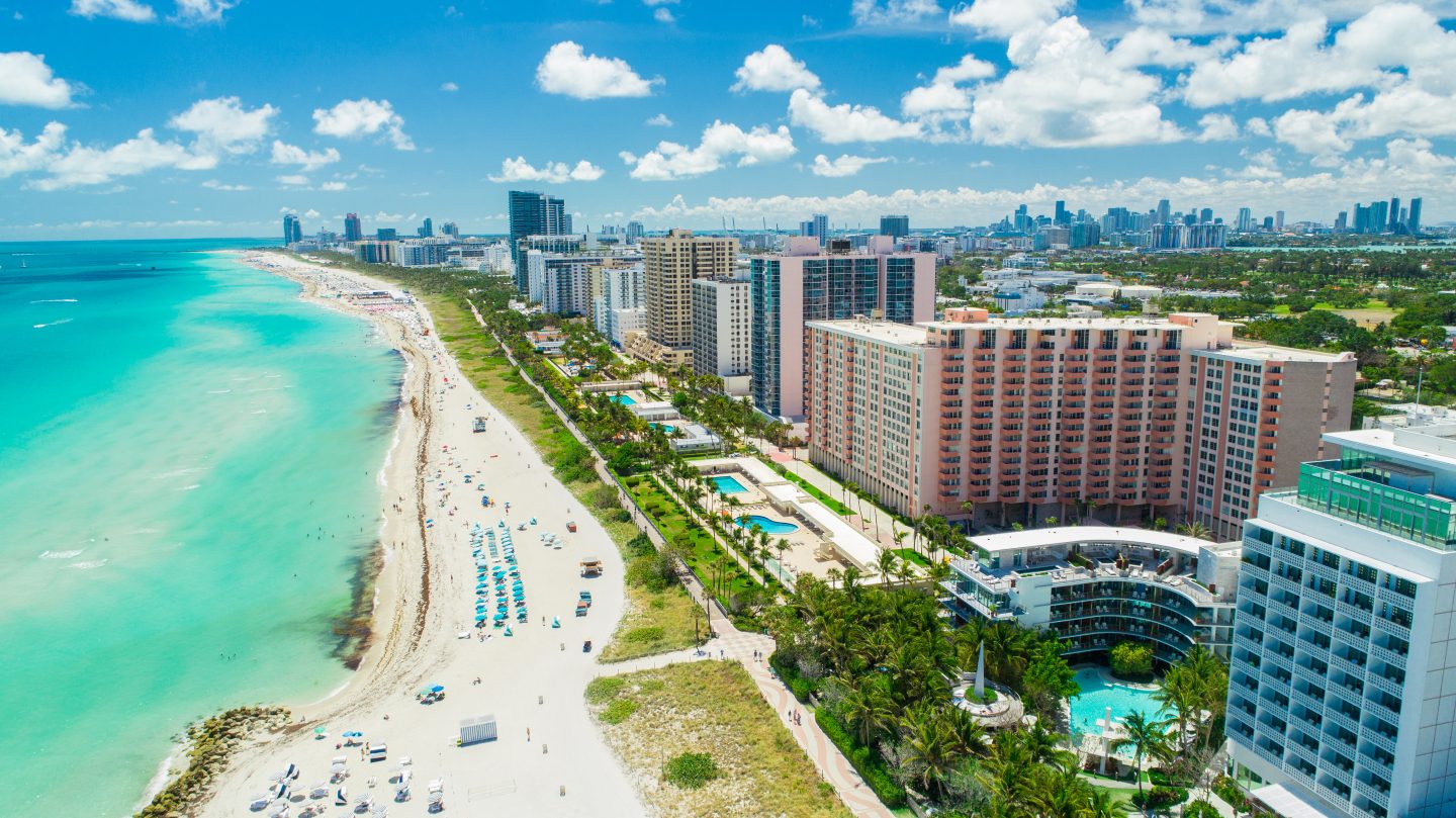 The Miami beach strip skyline of hotel towers along Miami beach on a hot sunny summer day. In the background is the city of Miami which is one of the cheapest summer destinations for 2023.