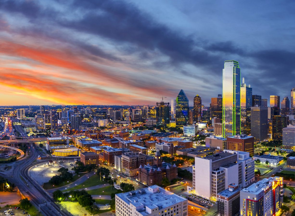 Dallas skyline at dusk with a partially cloudy sky and some of the buildings lit up neon green.