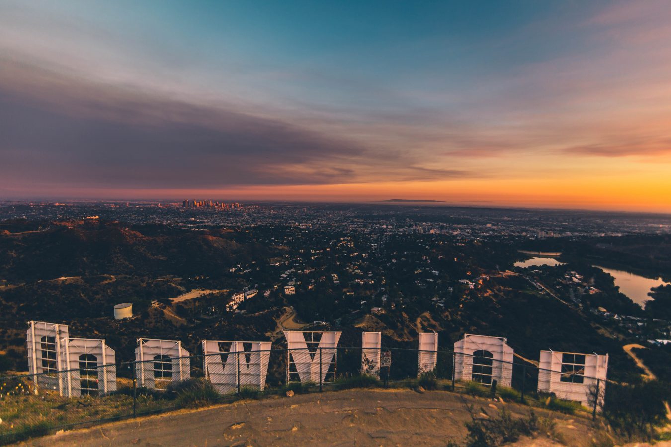 Overlooking Los Angeles from right behind the Hollywood sign at sunset. The sky is yellows, oranges, and blues, and the clouds are purples and pinks.