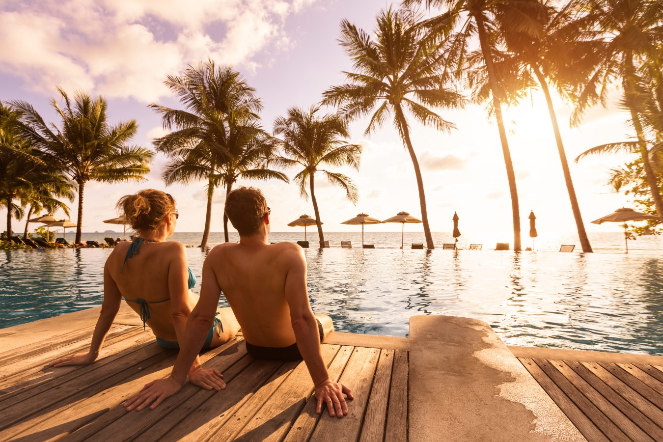 Couple enjoying beach vacation at tropical resort with swimming pool and coconut palm trees near the coast with beautiful landscape at sunset.