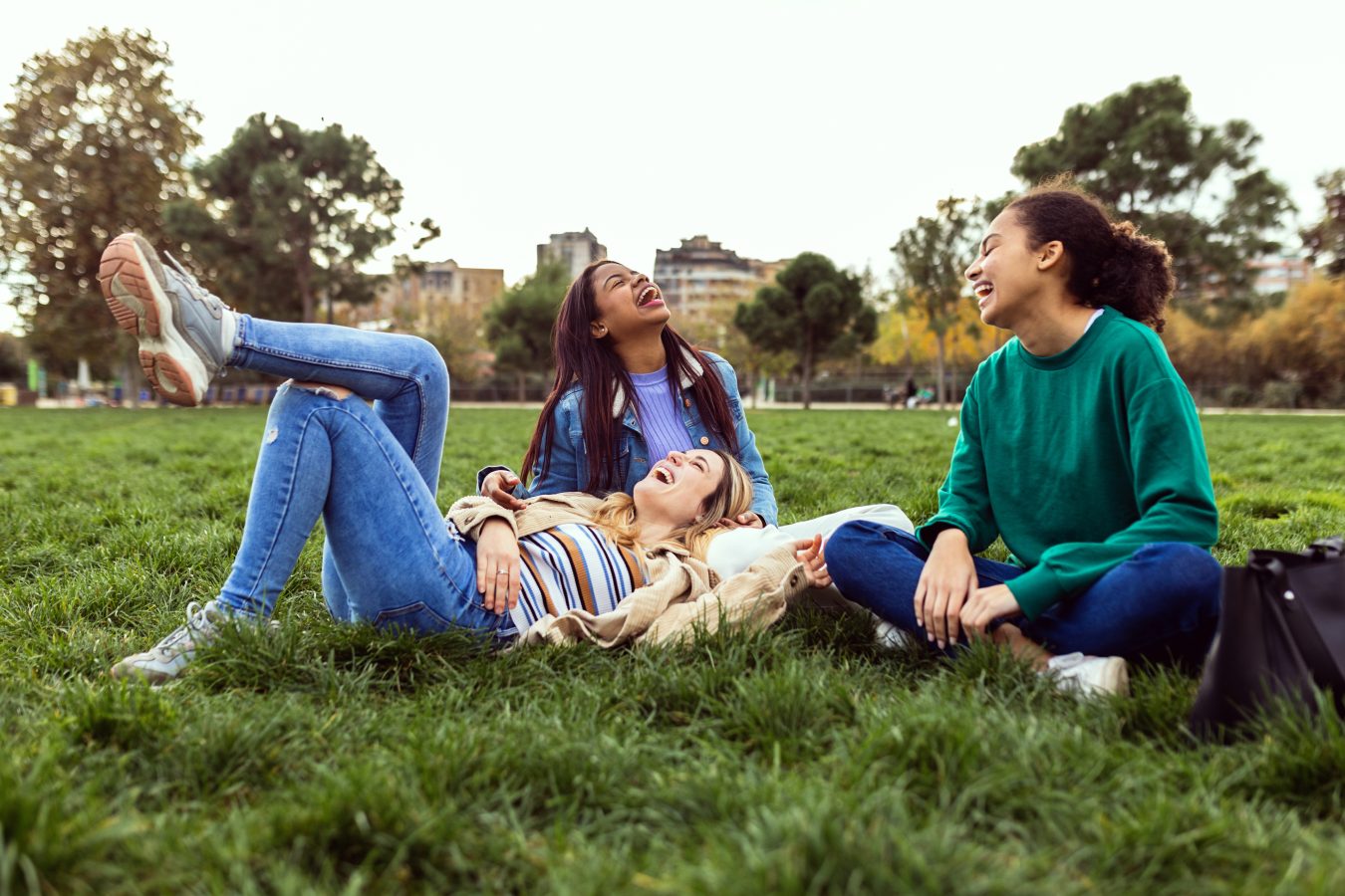 Three happy young women having fun sitting on green grass at college campus.