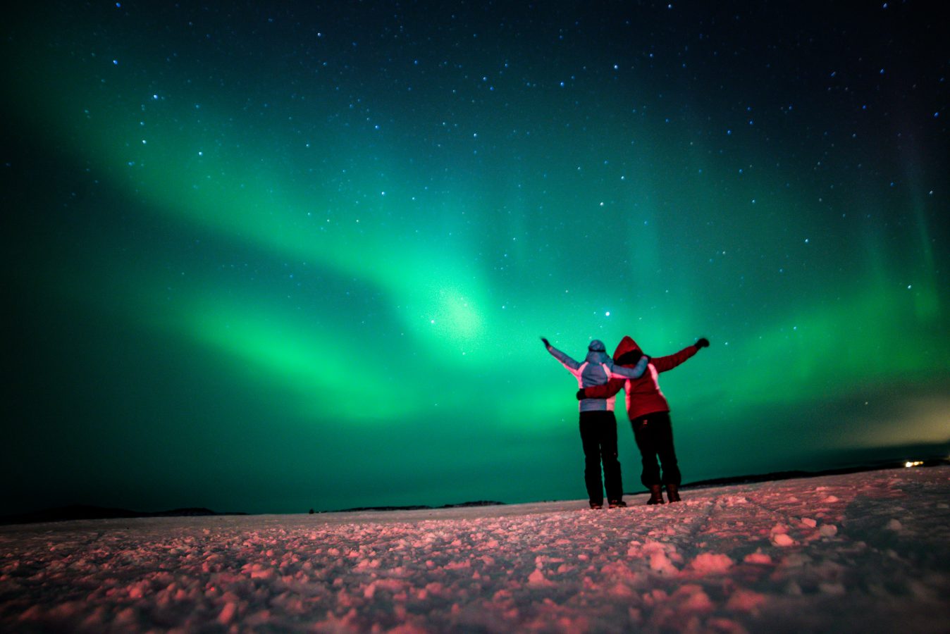 Two friends pose for a photo under the green Northern Lights. Seeing the Aurora Borealis is a cool winter getaway in Canada to go on.